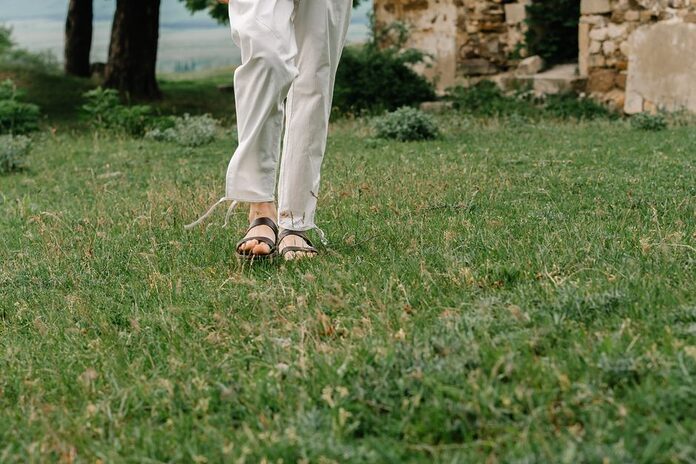 sandales-tendance-ete-2026-mode-sante-pieds A person walking on green grass wearing white pants and sandals, a relaxed outdoor scene.